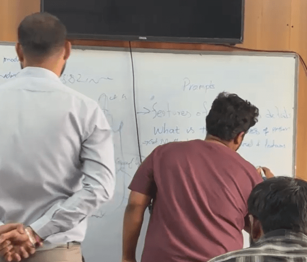 Ascent student standing at the classroom board writing and explaining feedback points while classmates watch, during a training session in the week of August 17–23, 2026